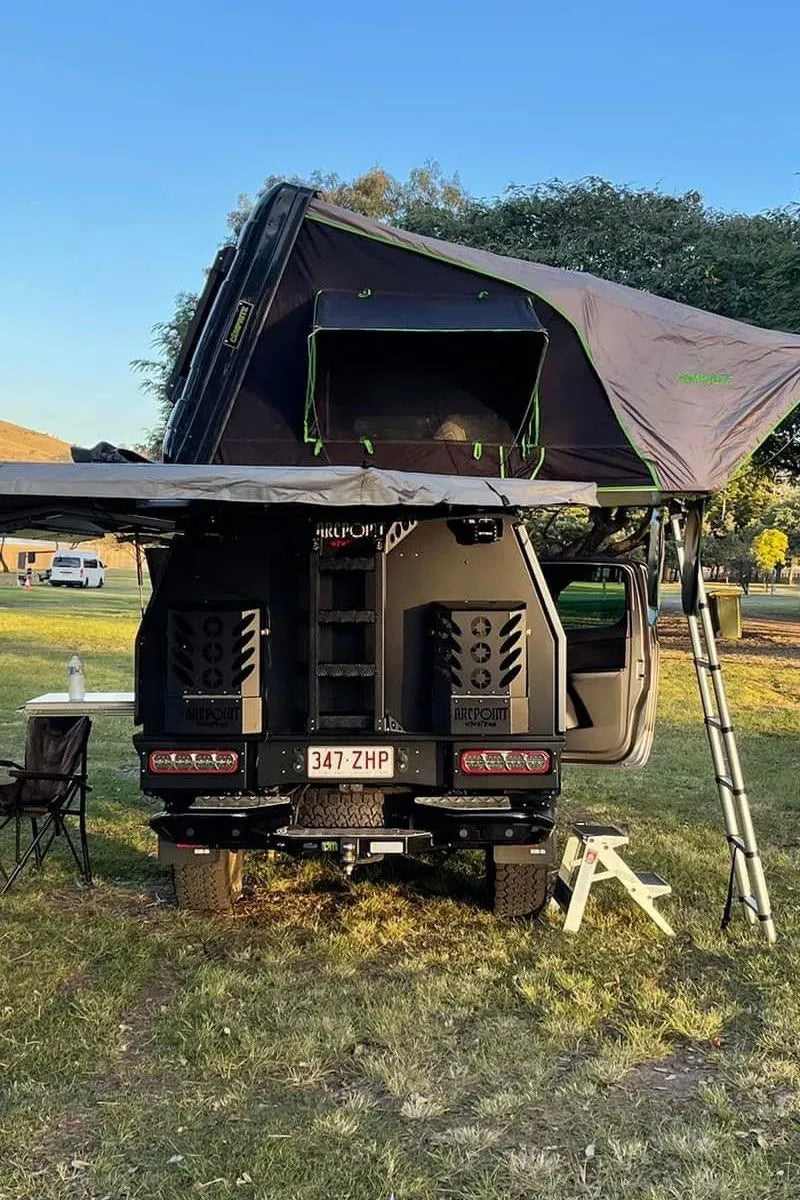 A rear view of a black and olive green camper van with a rooftop tent and awning set up outdoors on grassy ground under a blue sky.