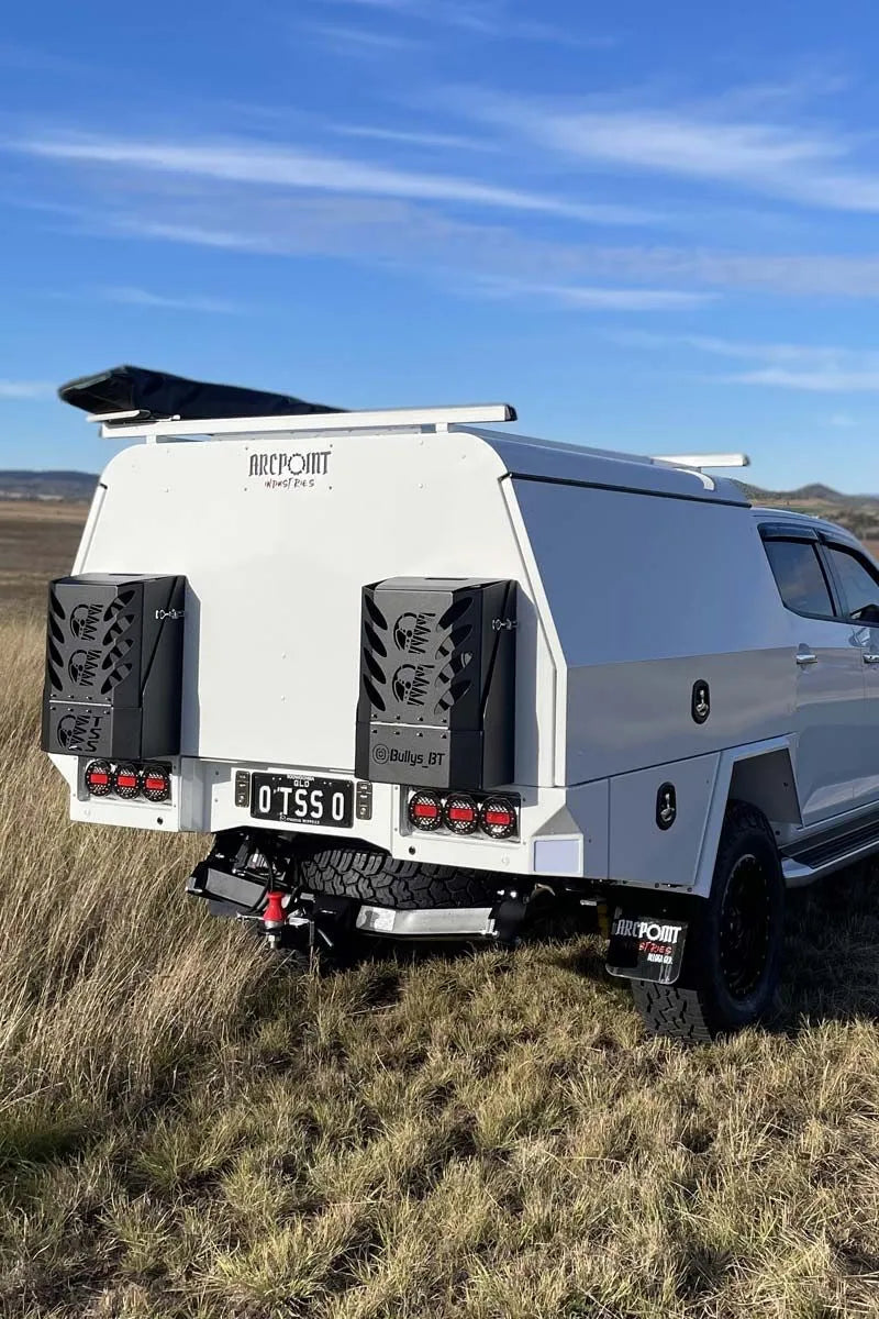 A white off-road camper trailer attached to a white pickup truck, parked in a grassy field under a blue sky. The rear of the trailer features black side-mounted storage units, a license plate, and the brand name 'ATKPOIN' printed on the top panel.