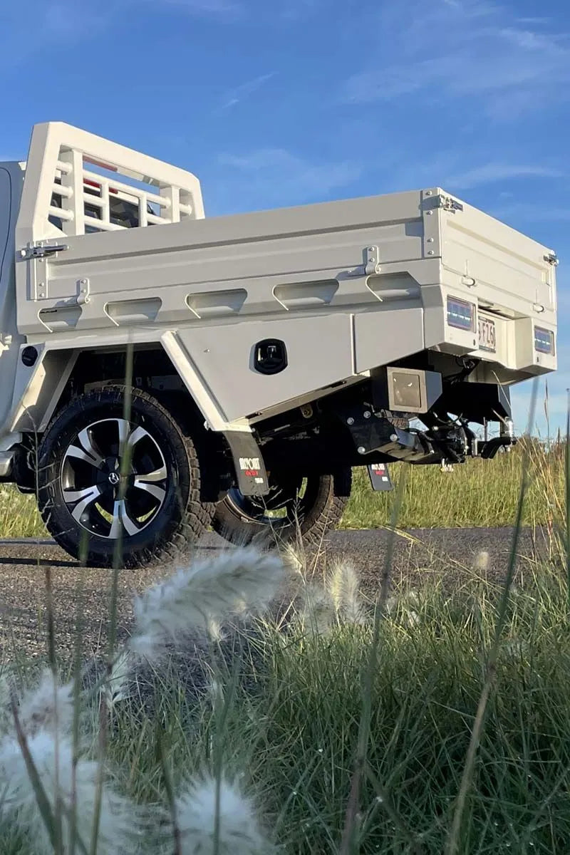 A light beige utility truck bed with black tires and a black undercarriage, parked outdoors on a paved path with tall grass in the foreground and a blue sky behind.