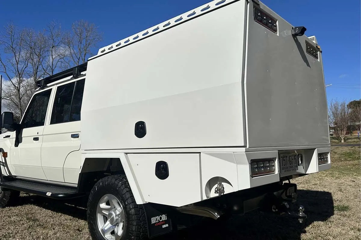 A white off-road vehicle with a large white hard body shell or camper-style box mounted over the bed, photographed outdoors on grass under a blue sky. No people or animals are present.