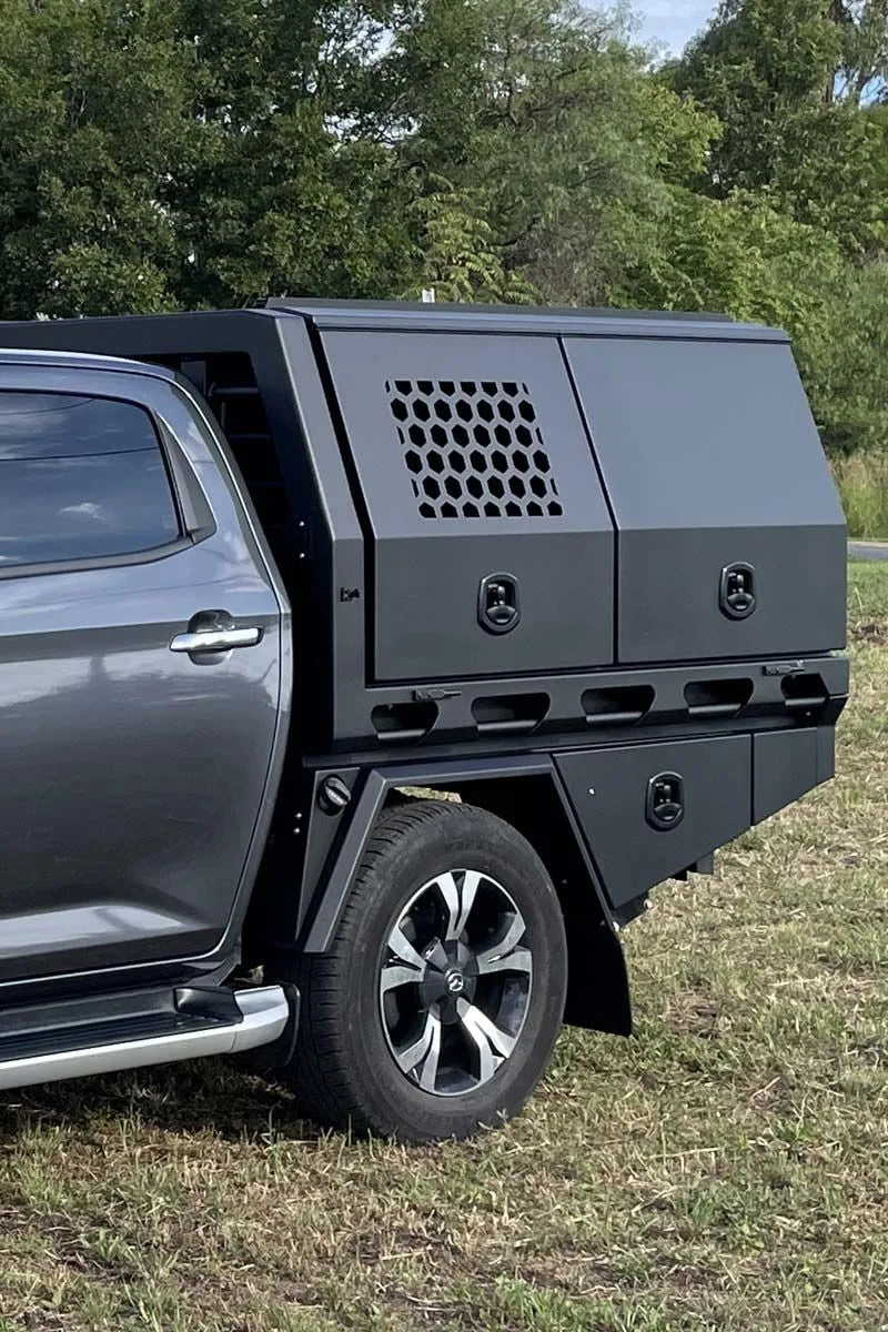 A dark gray hardtop truck bed cover installed on a gray pickup truck, parked outdoors on grass with trees in the background.