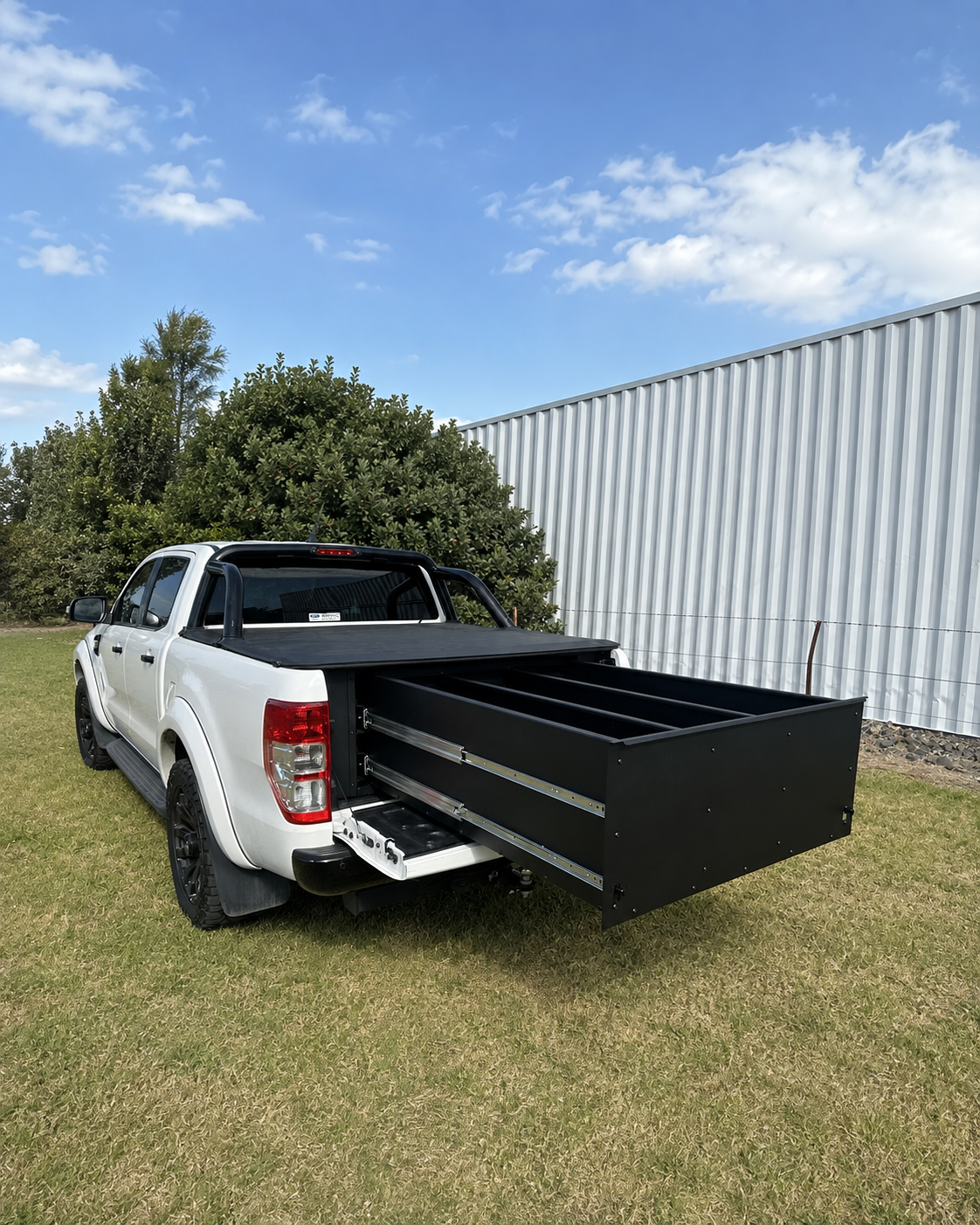 A white pickup truck parked on grass outdoors, with a large black metal utility bed or storage box extended from the bed area. Trees, a blue sky with clouds, and a corrugated metal building are visible in the background.